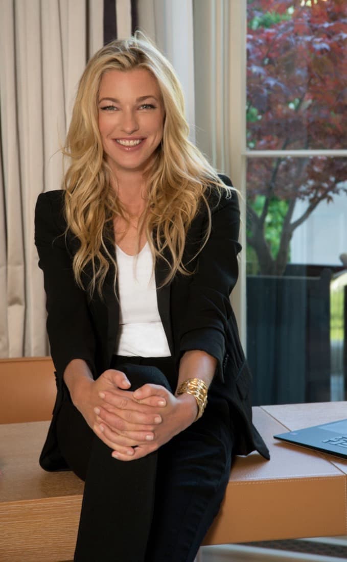 Nicole Junkermann smiling warmly, seated on a desk beside a window